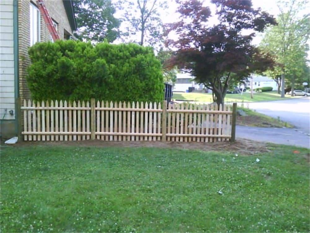 Wooden picket fence in front yard with green lawn and shrubs, near a building and street.
