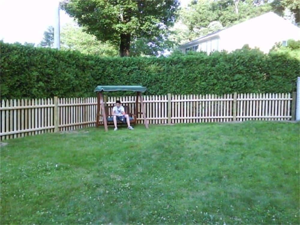 Person seated on a swing set in a grassy yard, enclosed by a wooden fence and hedge.