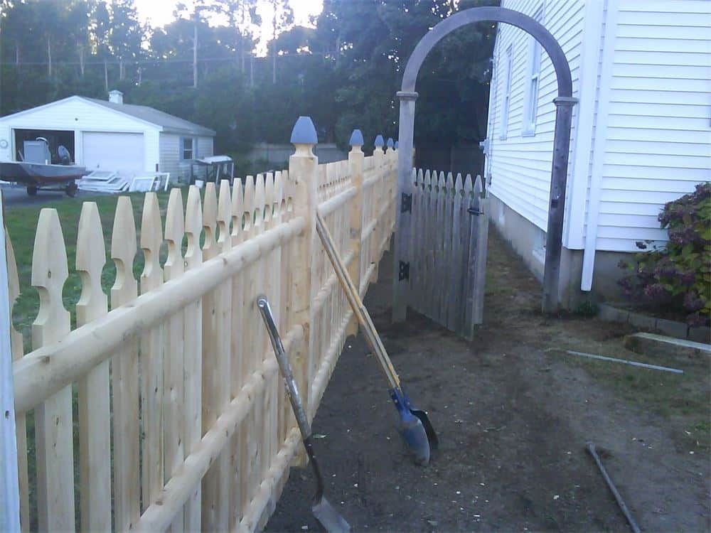 Wooden picket fence with a gate and archway next to a white house. Two tools lean against the fence.