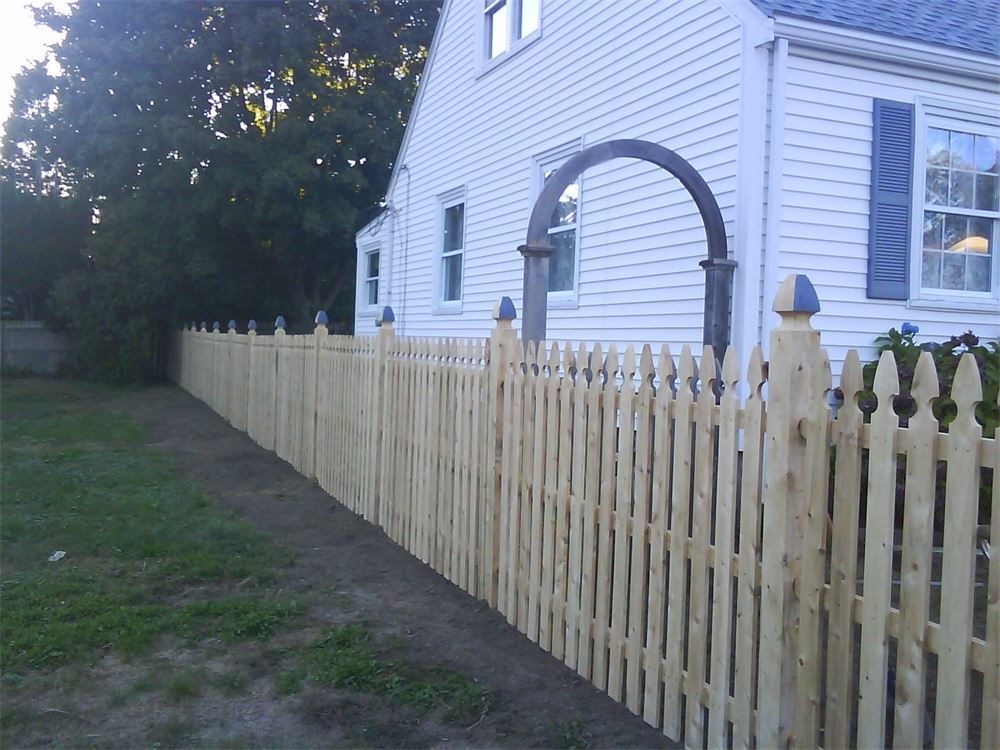 Wooden picket fence in front of a white house with blue shutters, a dark arch, and green grass.
