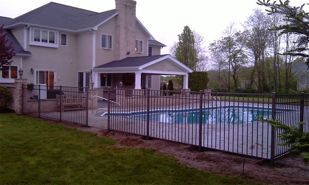 Backyard with a swimming pool enclosed by a black metal fence, and a large house with a covered patio.
