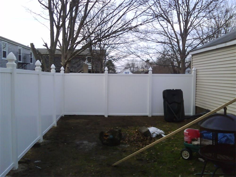White vinyl fence encloses a small, bare backyard with a fire pit and gardening tools. Overcast sky.