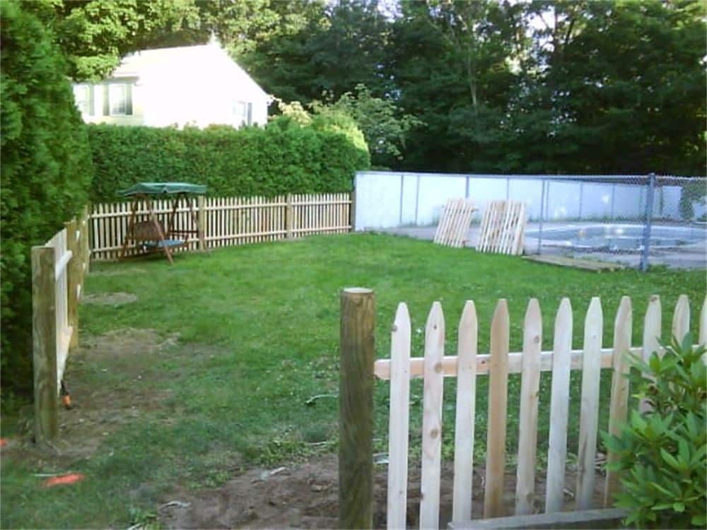Wooden picket fence surrounding a grassy yard with swing set, green hedge, and house in the background.