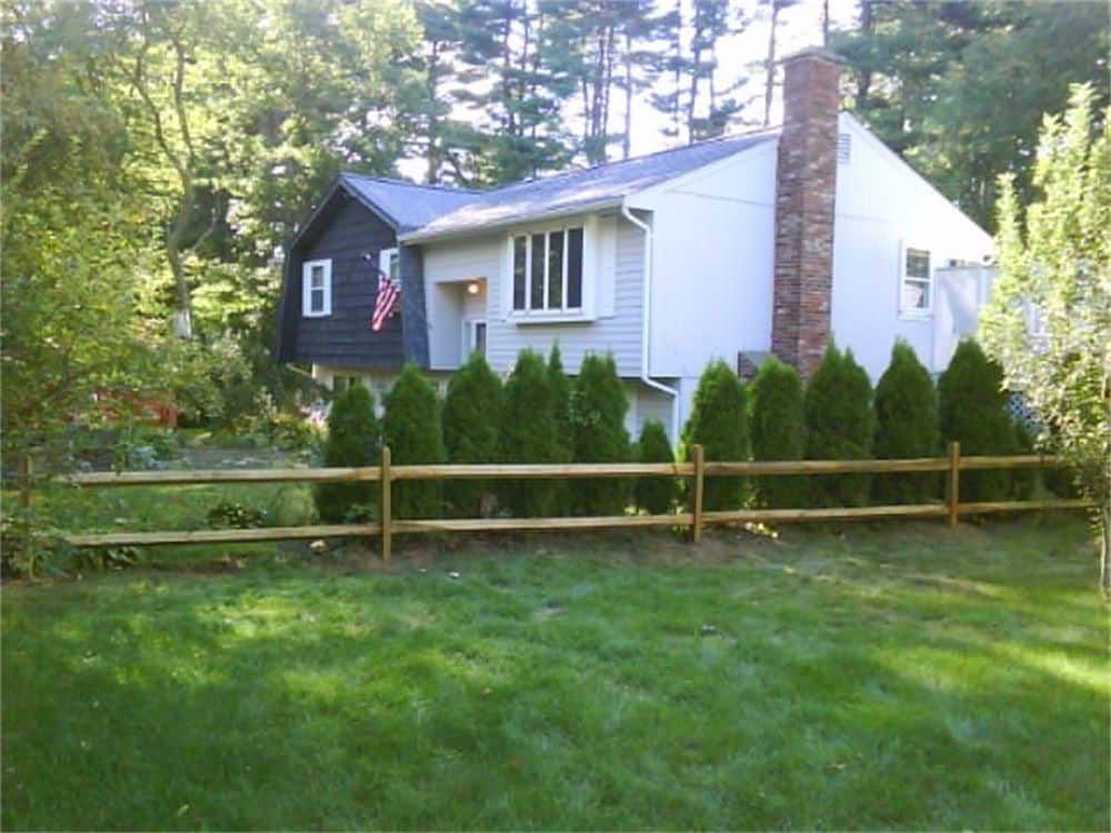 House with light gray siding, wooden fence, and evergreen trees. American flag hanging.