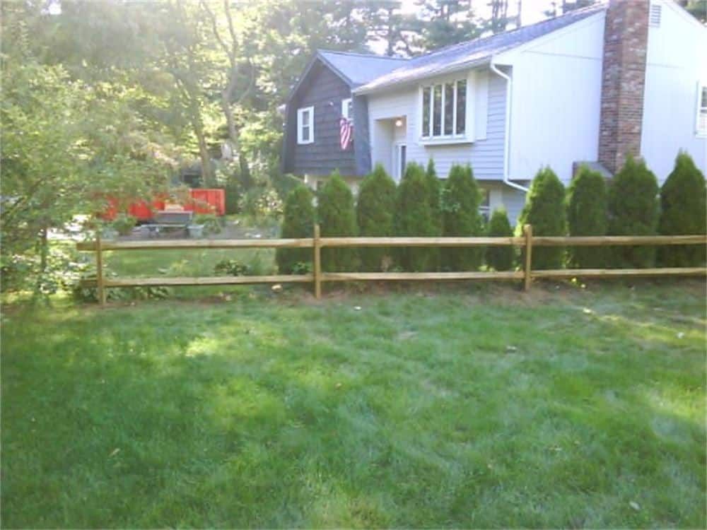 Wooden split-rail fence in front of a house with green grass and evergreen trees.