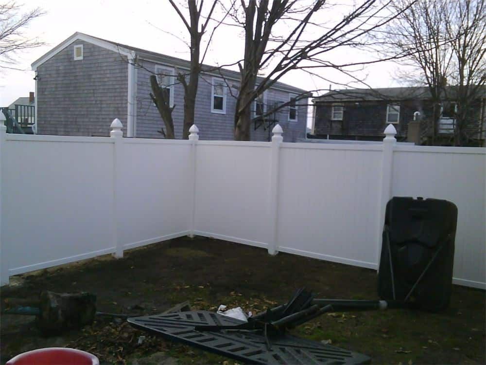 White vinyl fence encloses a backyard; houses and bare trees in the background.