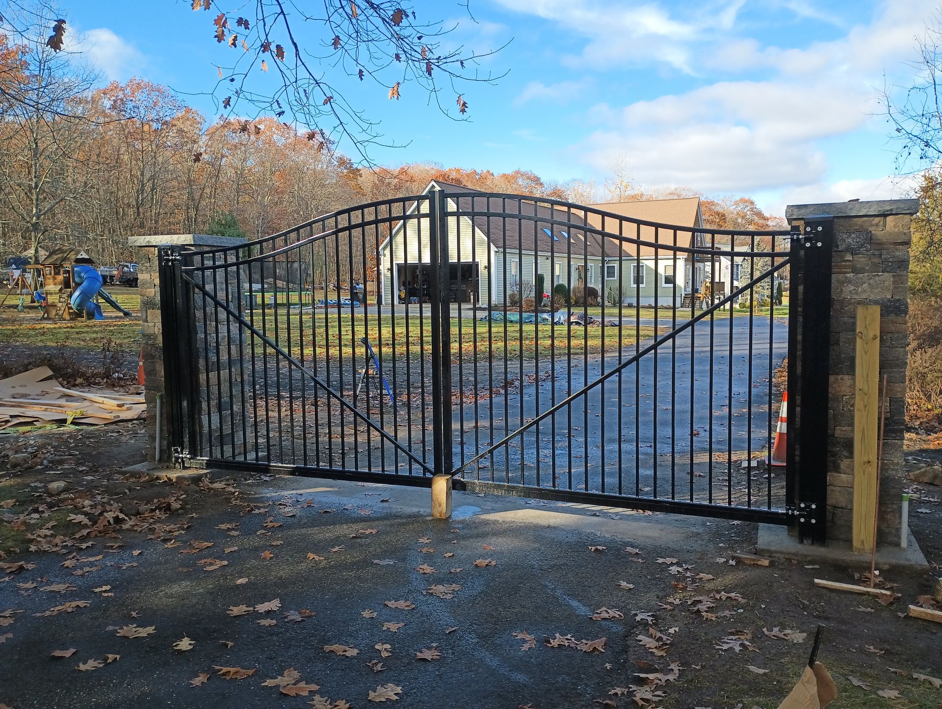 Black metal driveway gate between stone pillars, leading to a house in a wooded area.