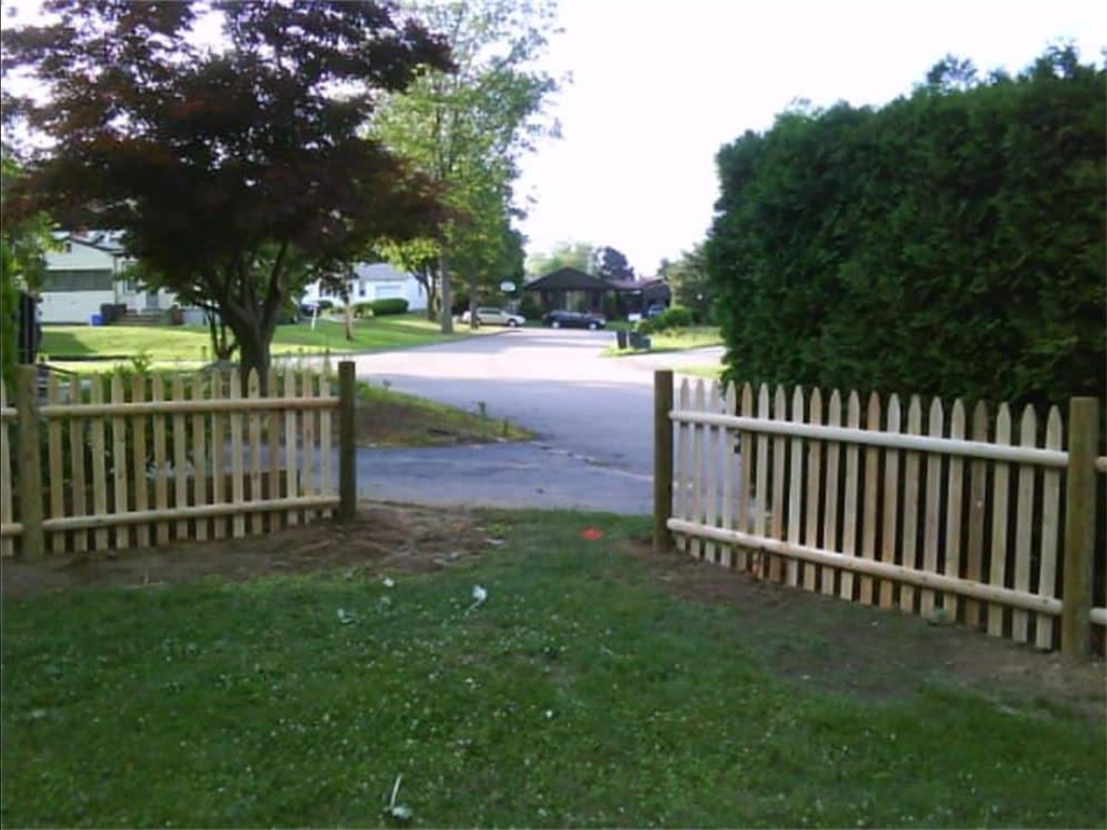 Open picket fence gate leading to a residential street. Green grass and trees frame the view.