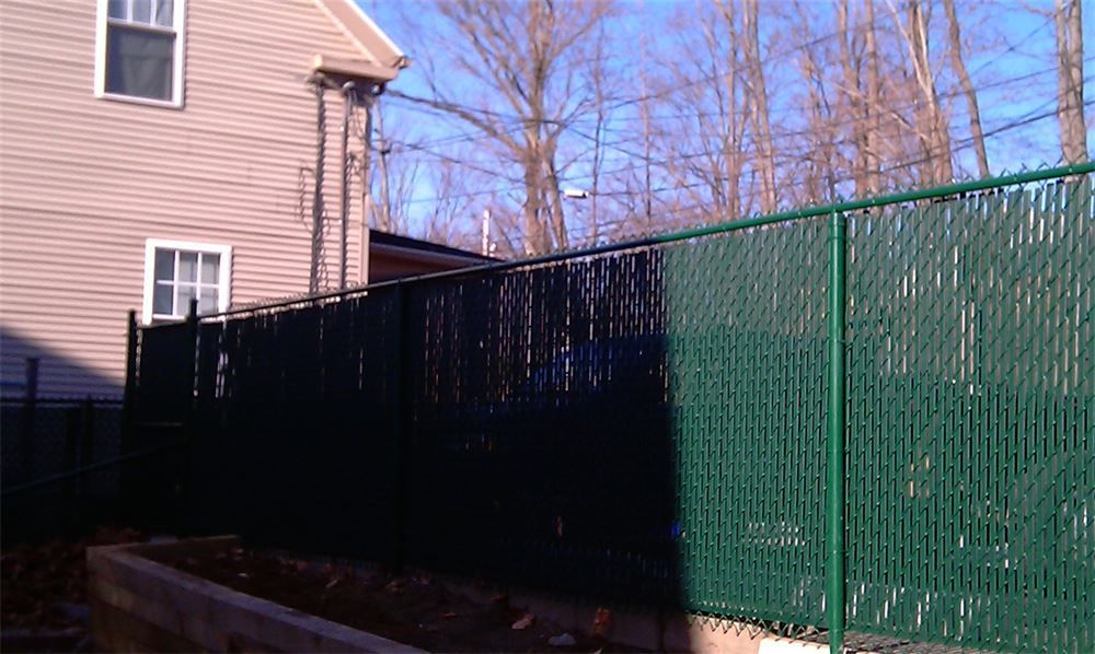 Green chain-link fence with privacy slats along the side of a beige house with a clear blue sky in the background.