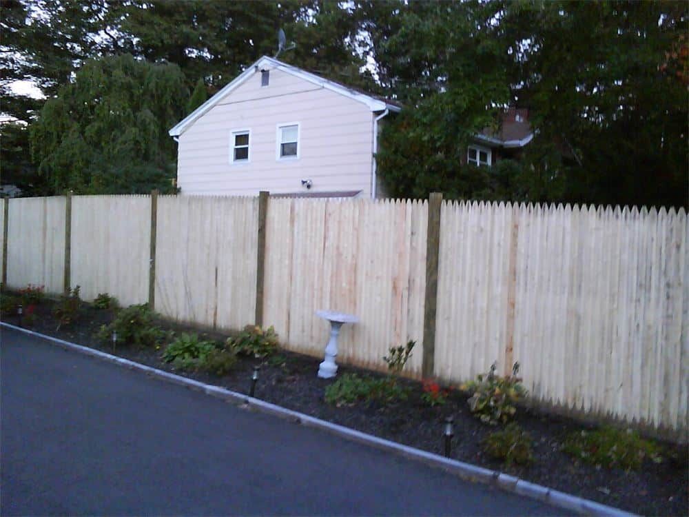 Wooden fence in front of a light-colored house; bird bath in front of the fence, plants along the road.