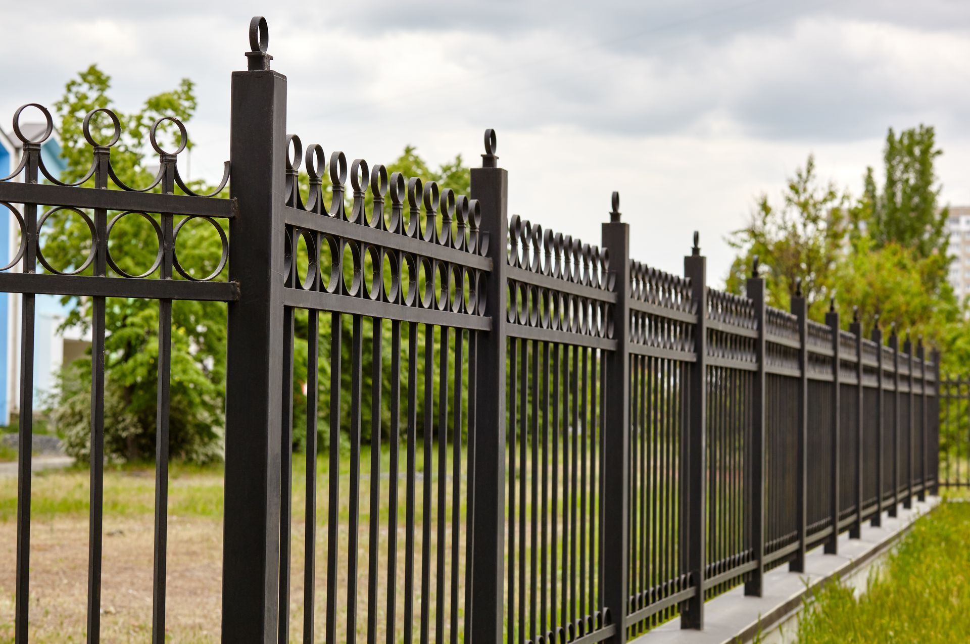 Black metal fence along a grassy area, trees in the background, cloudy sky.