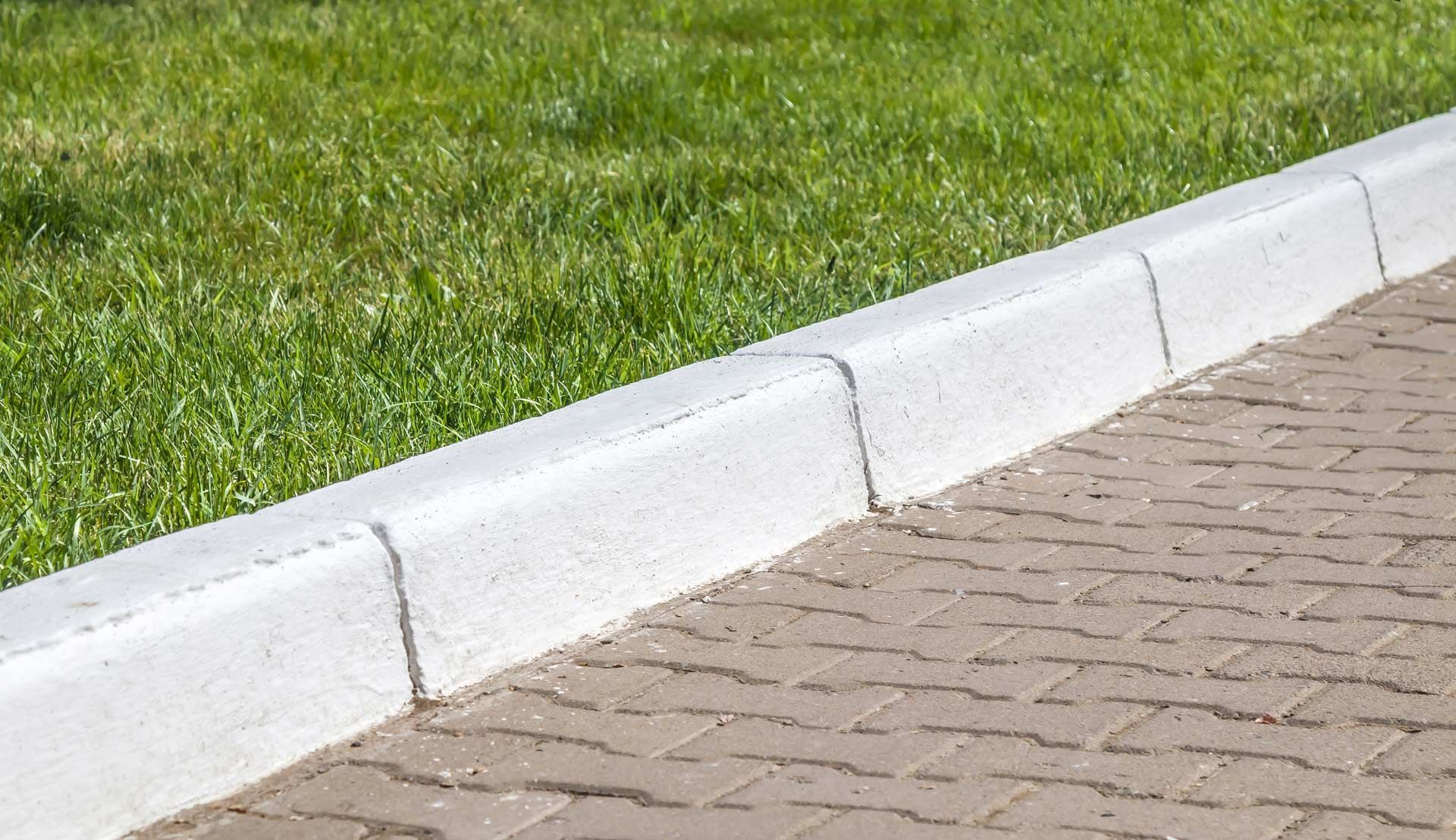 White concrete curb separating a brick paved walkway from green grass.
