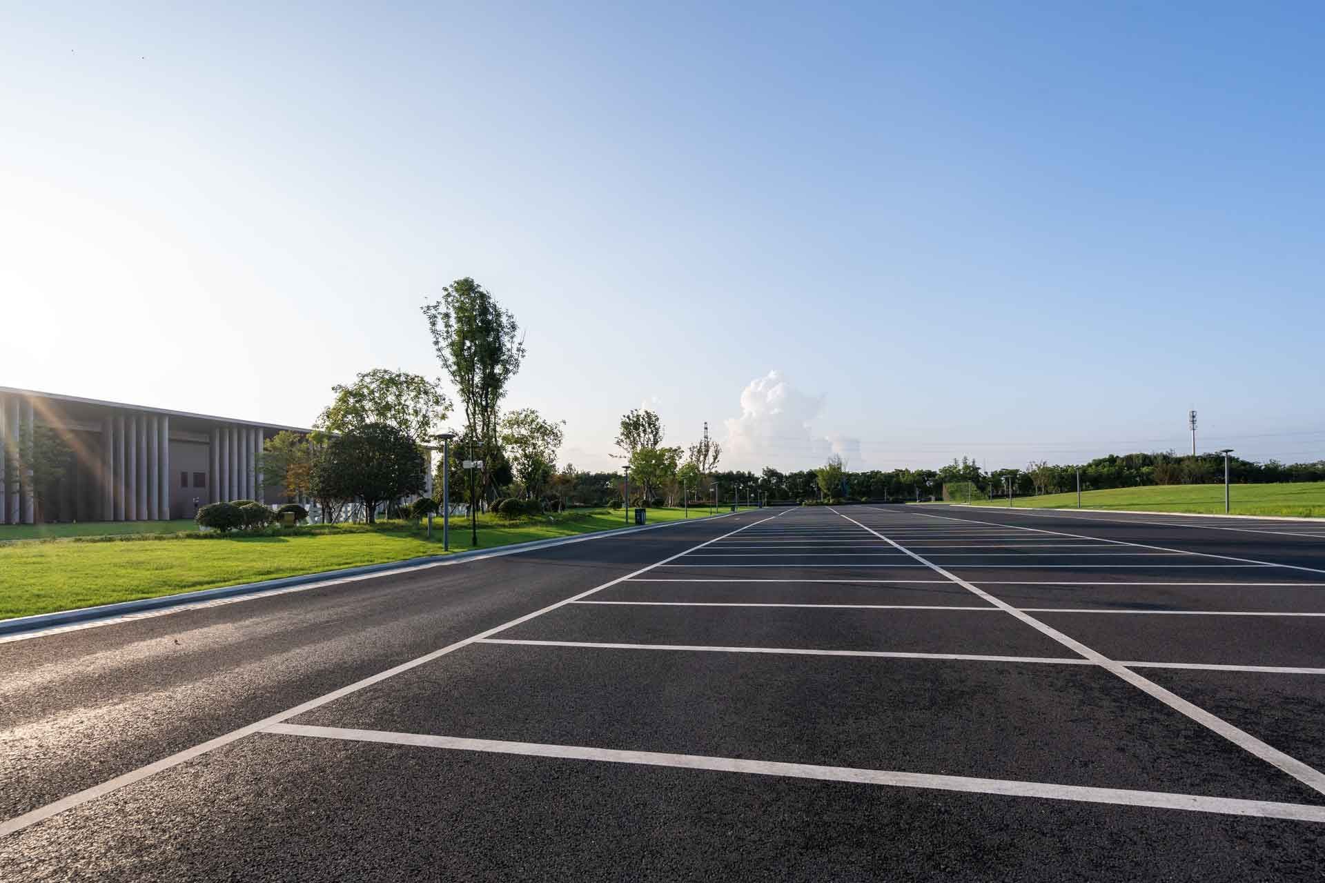 Empty asphalt parking lot with white lines, grass, and trees under a blue sky.