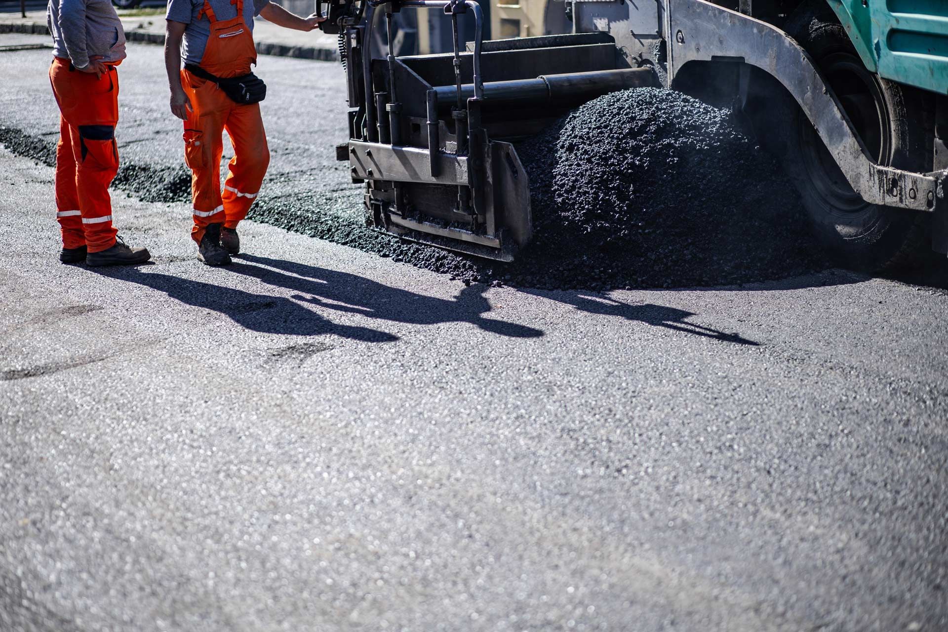 Two road workers in orange coveralls near an asphalt paver, laying new asphalt on a road.