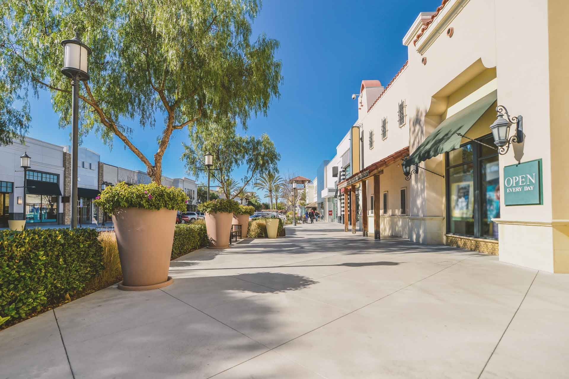 Outdoor shopping center with beige buildings, lush trees, and a bright blue sky.