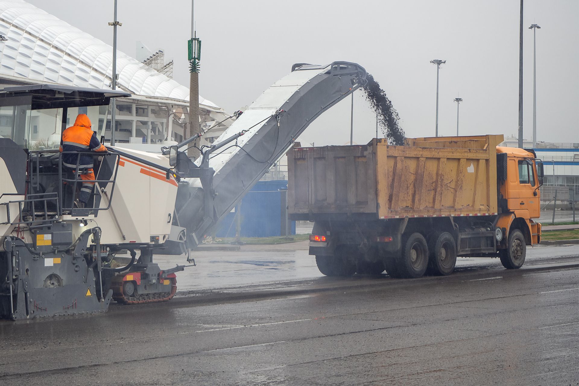 Road milling machine loading asphalt into an orange dump truck on a wet road.