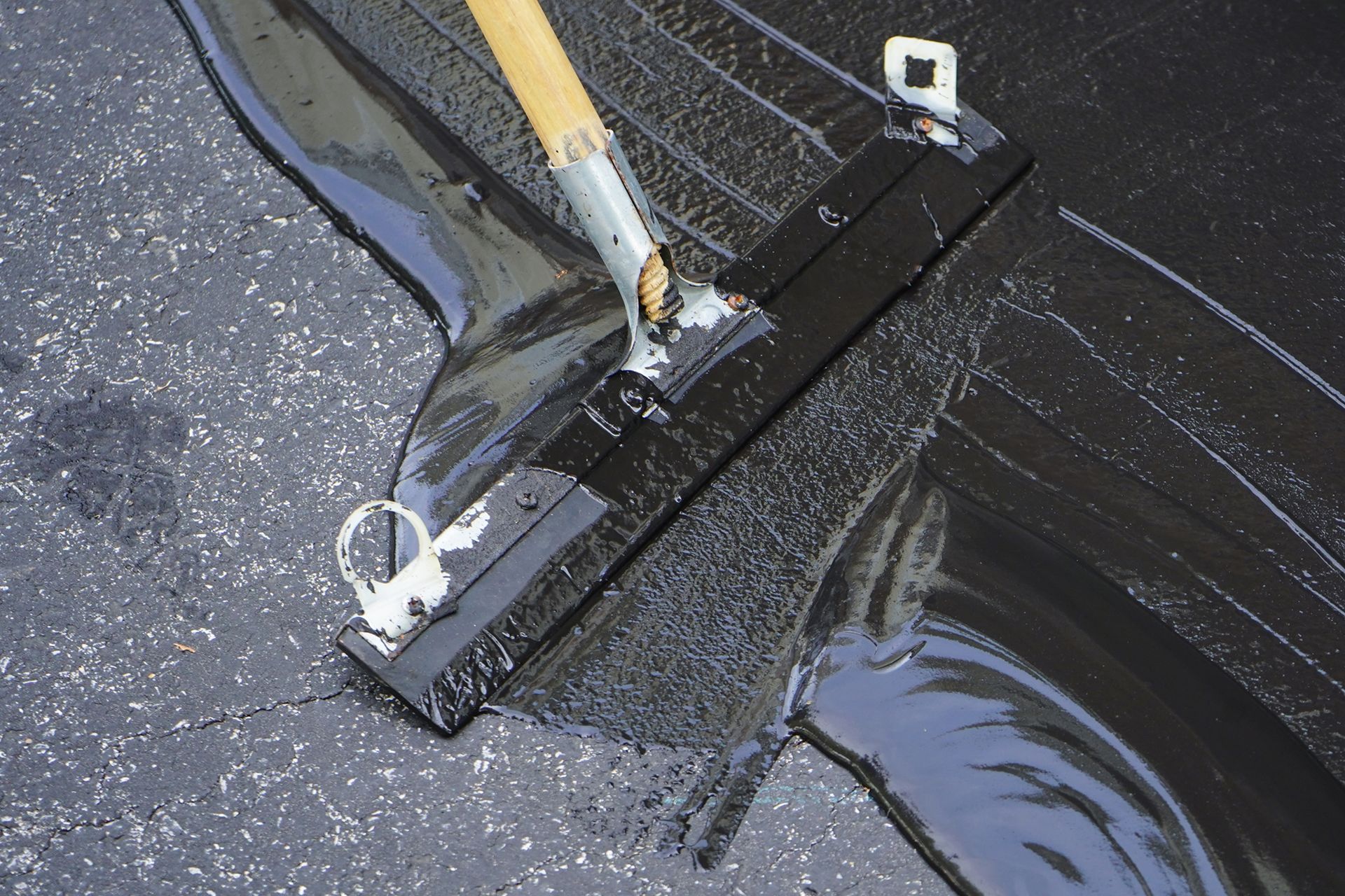 Person using a squeegee to apply black asphalt sealer to a cracked parking lot.