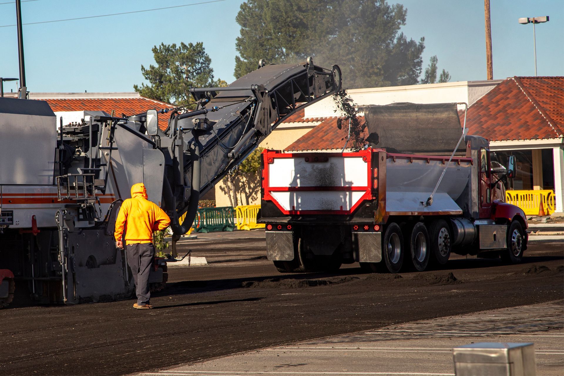 A road milling machine loading asphalt into a dump truck. Worker in yellow vest watches.
