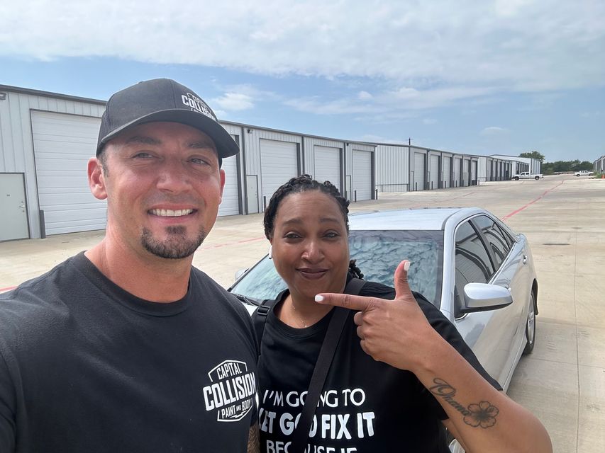 Two people smiling, standing next to a silver car in front of storage units on a sunny day.