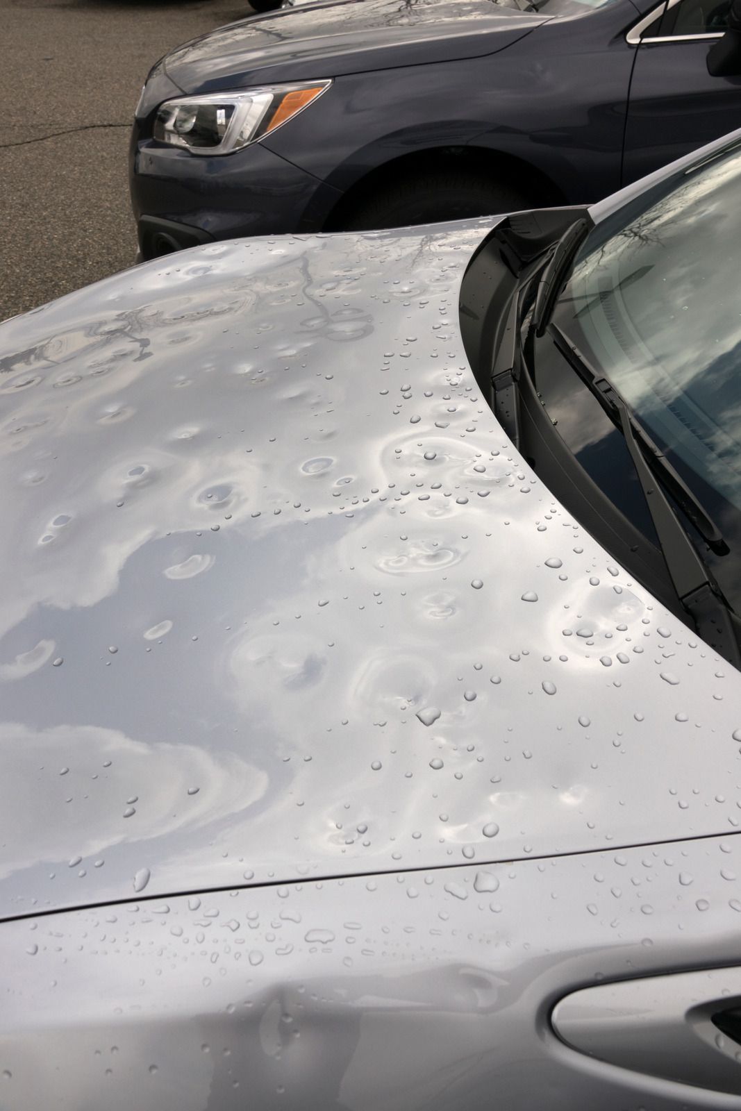 A silver car with hail damage on the hood is parked next to a blue car.