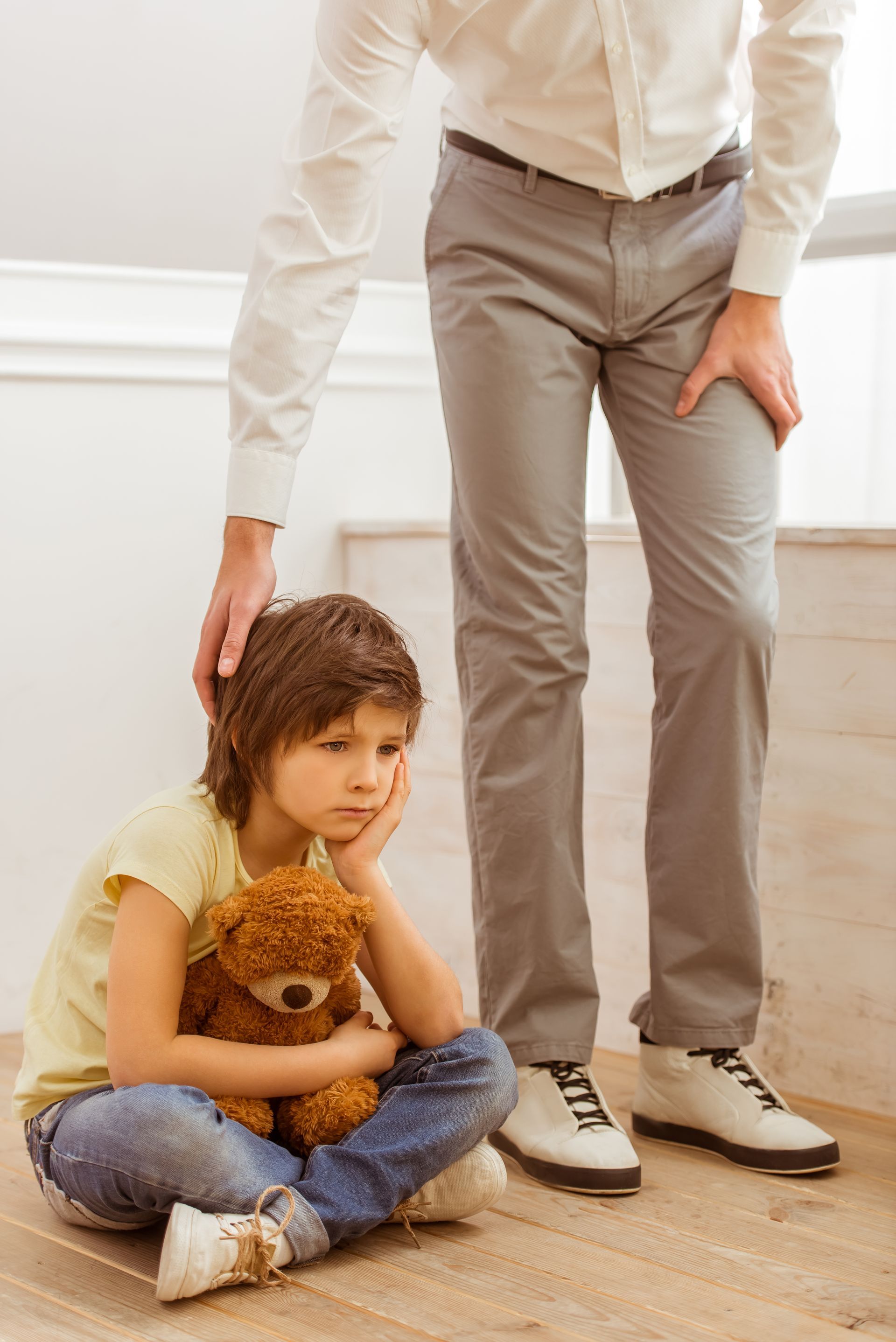 Man comforts a child sitting on the floor, holding a teddy bear; indoors.