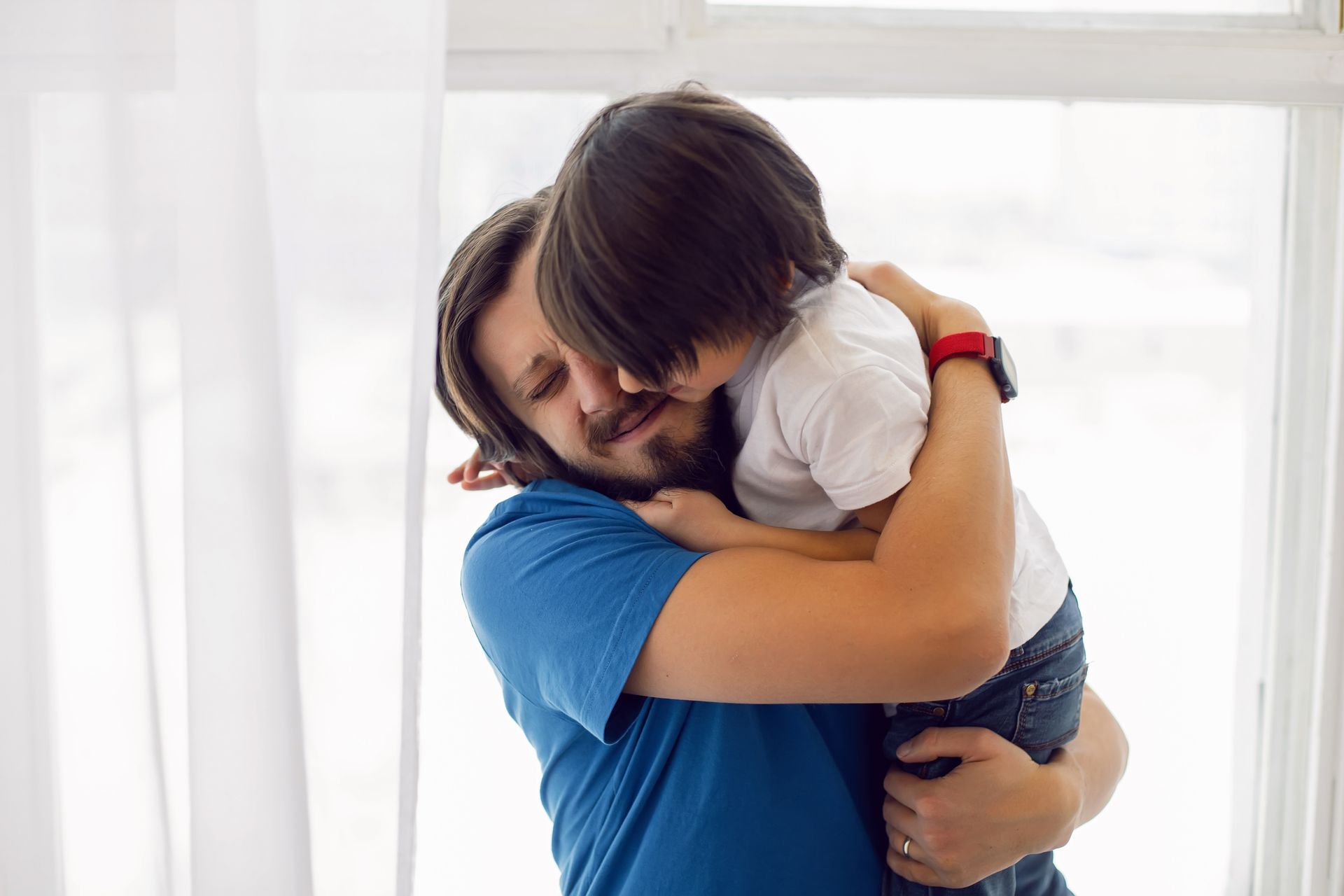 Man hugging a child by a window; both are embracing each other.
