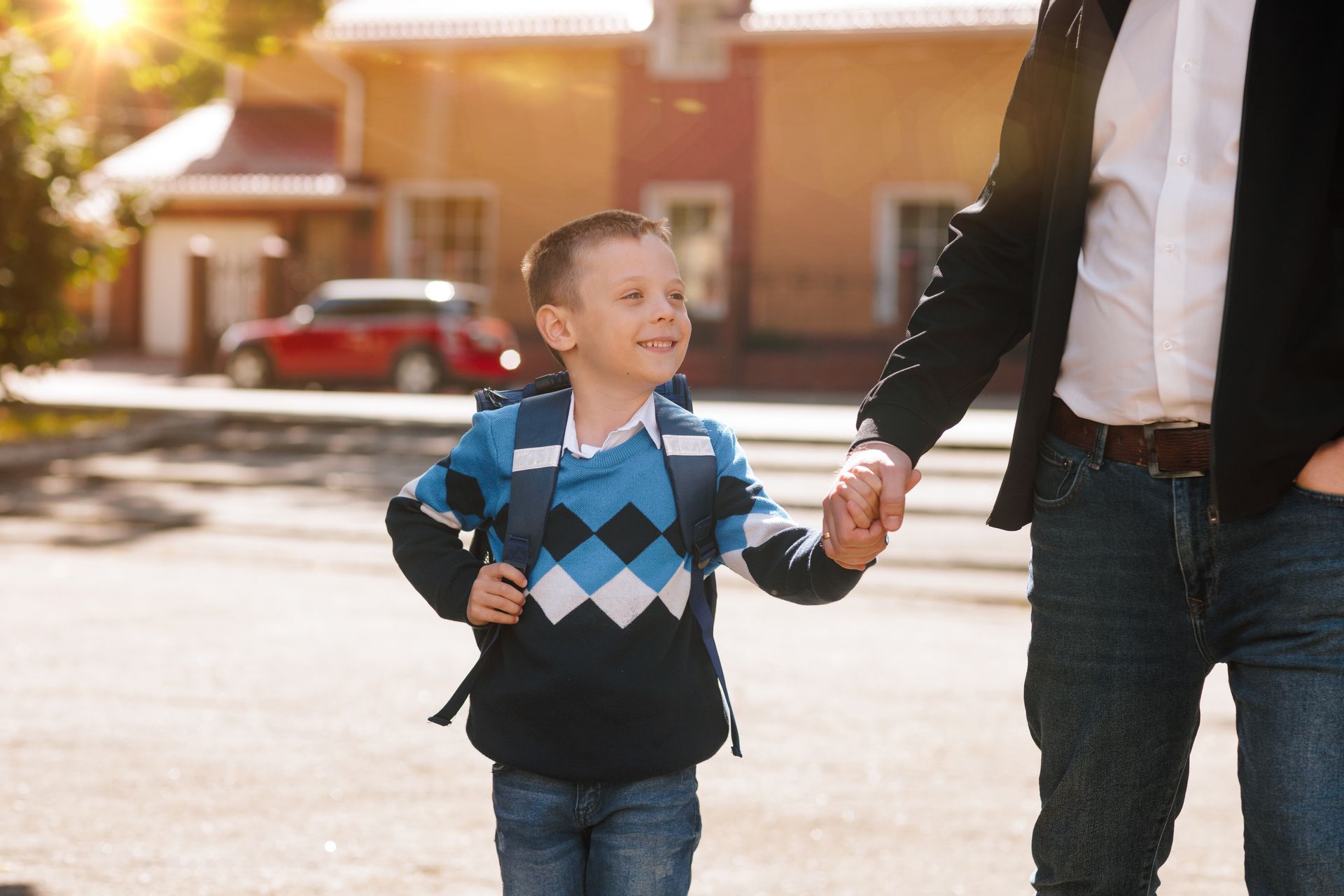 Boy with backpack holding hands with an adult, walking on a street toward a building on a sunny day.
