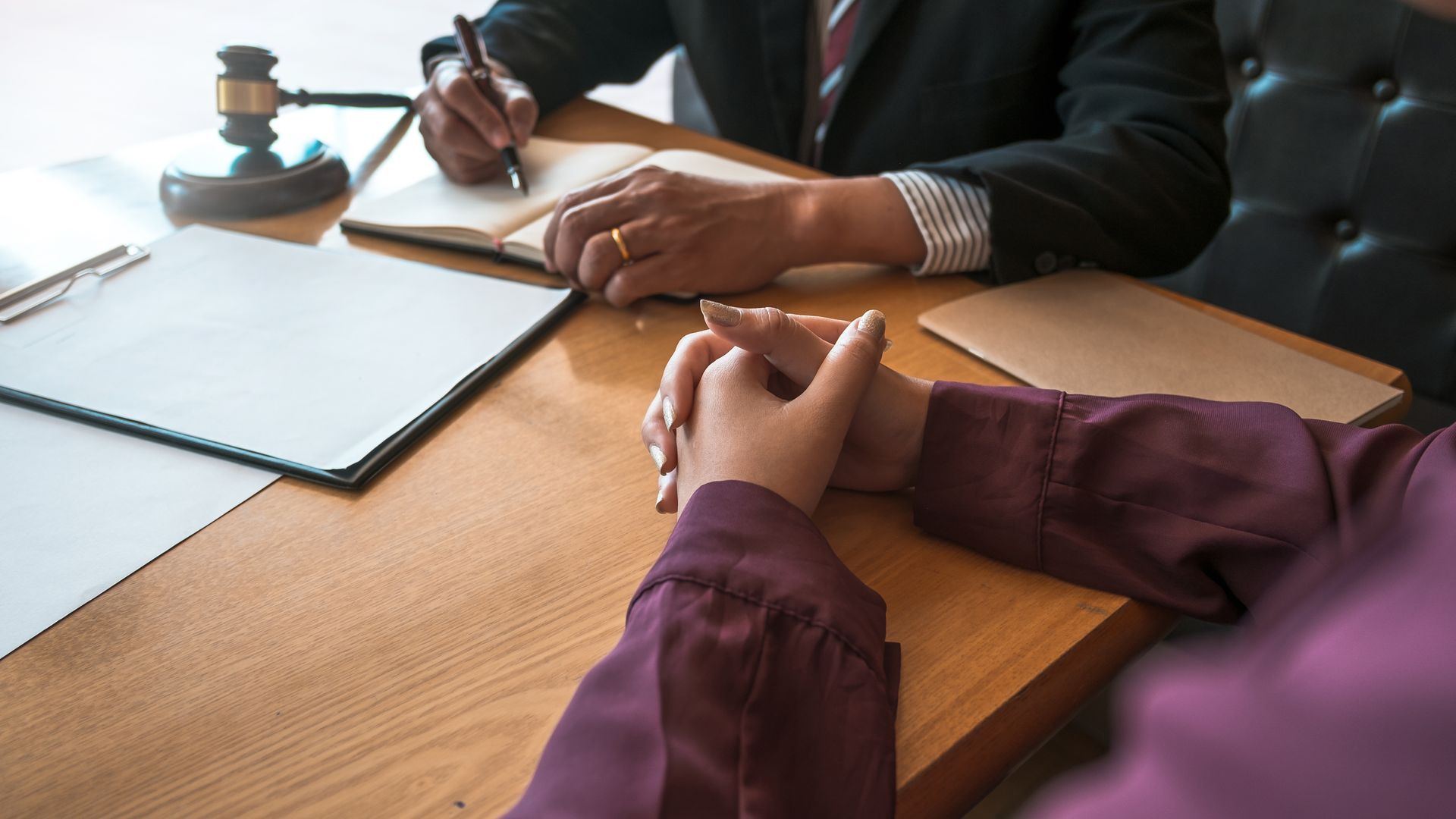 Hands clasped on a table across from a person taking notes, legal papers, and a gavel.