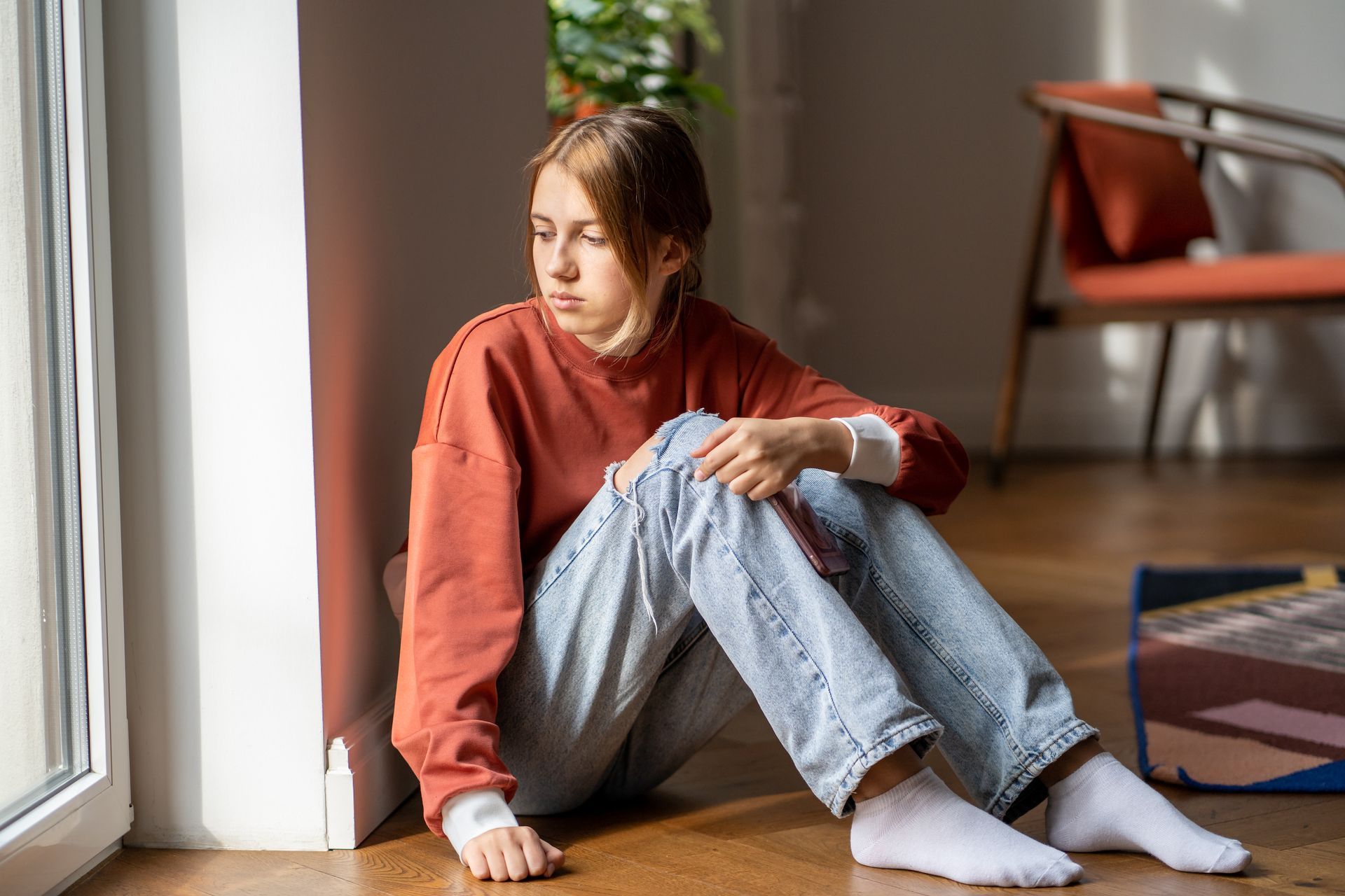 A person in a red sweater and jeans sits near a window, looking sad.