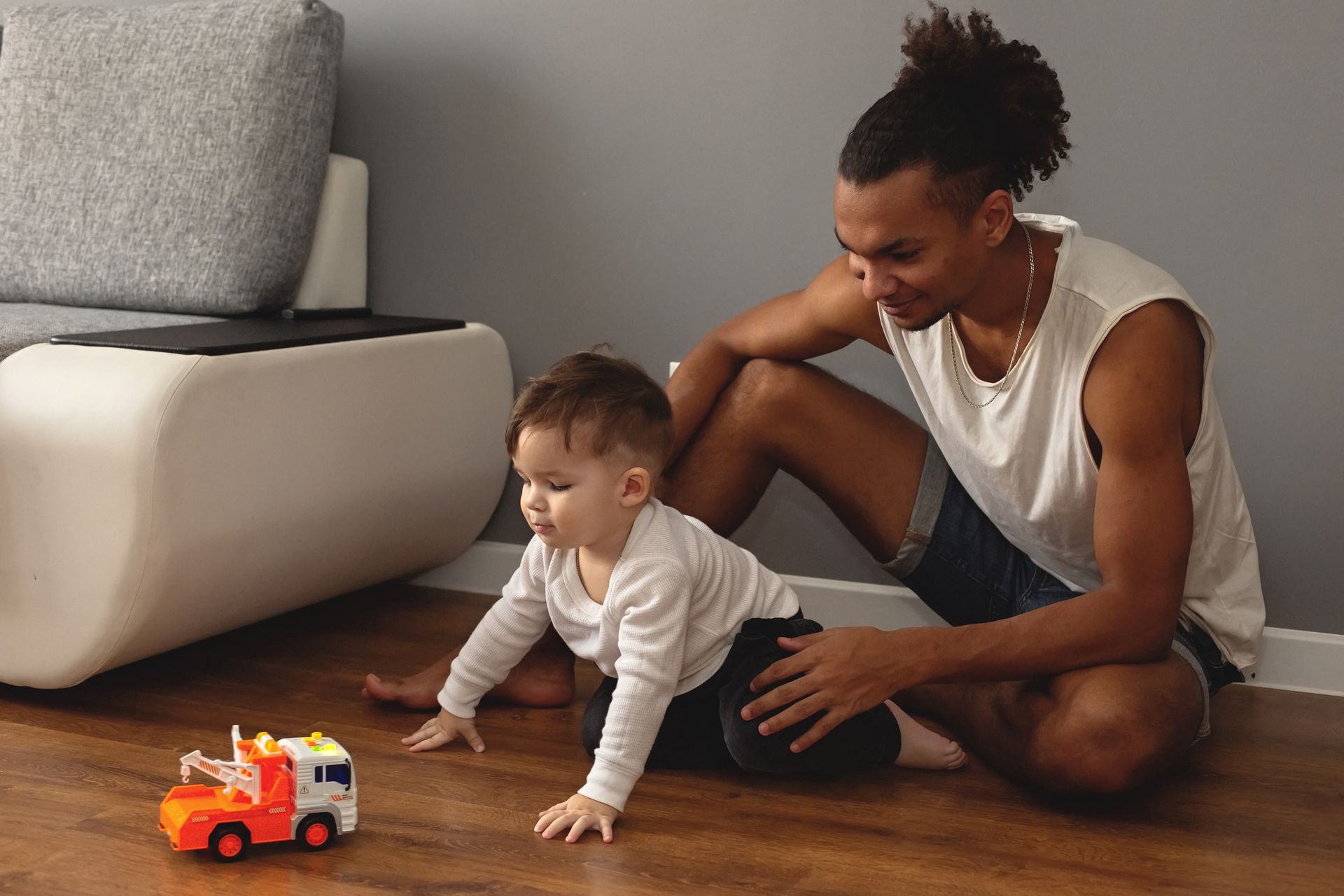 Man watching baby crawl towards a toy truck on a hardwood floor.