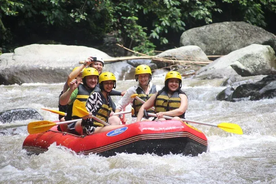 People rafting on a river, wearing helmets and life vests. Red raft, rocky riverbank, trees in the background.