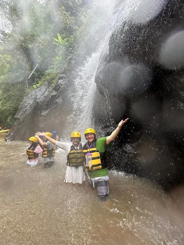 People in life vests pose near a waterfall, arms raised, smiling. Set in a river, the water cascades down a dark rock face.