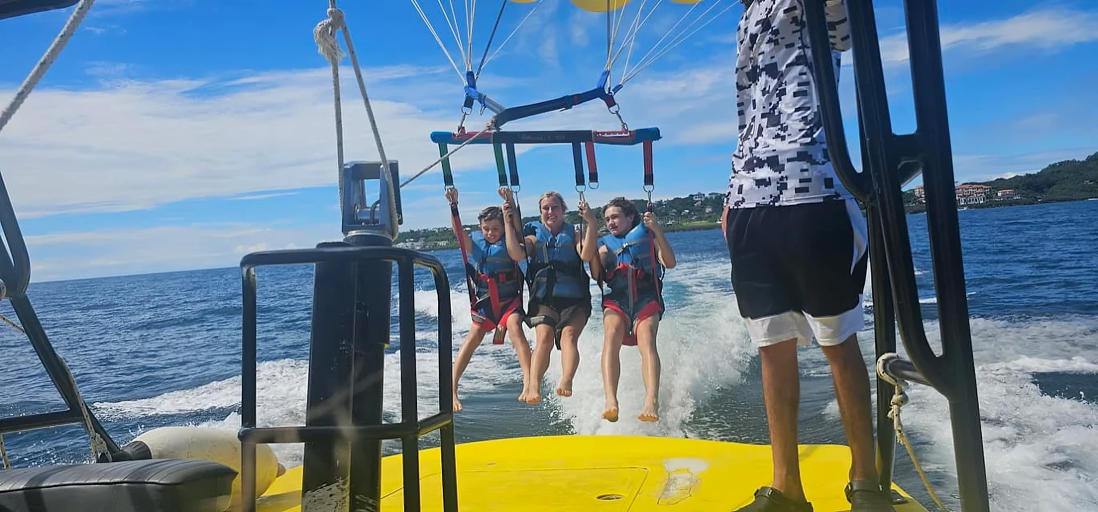 Three people parasailing, lifted from a yellow boat on a sunny day. They are wearing life vests and smiling as the boat moves forward.