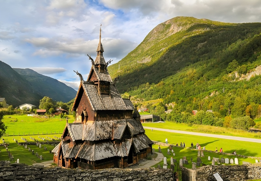 A wooden stave church with tiered roofs, set in a green valley with mountains in the background; graveyard in the foreground.
