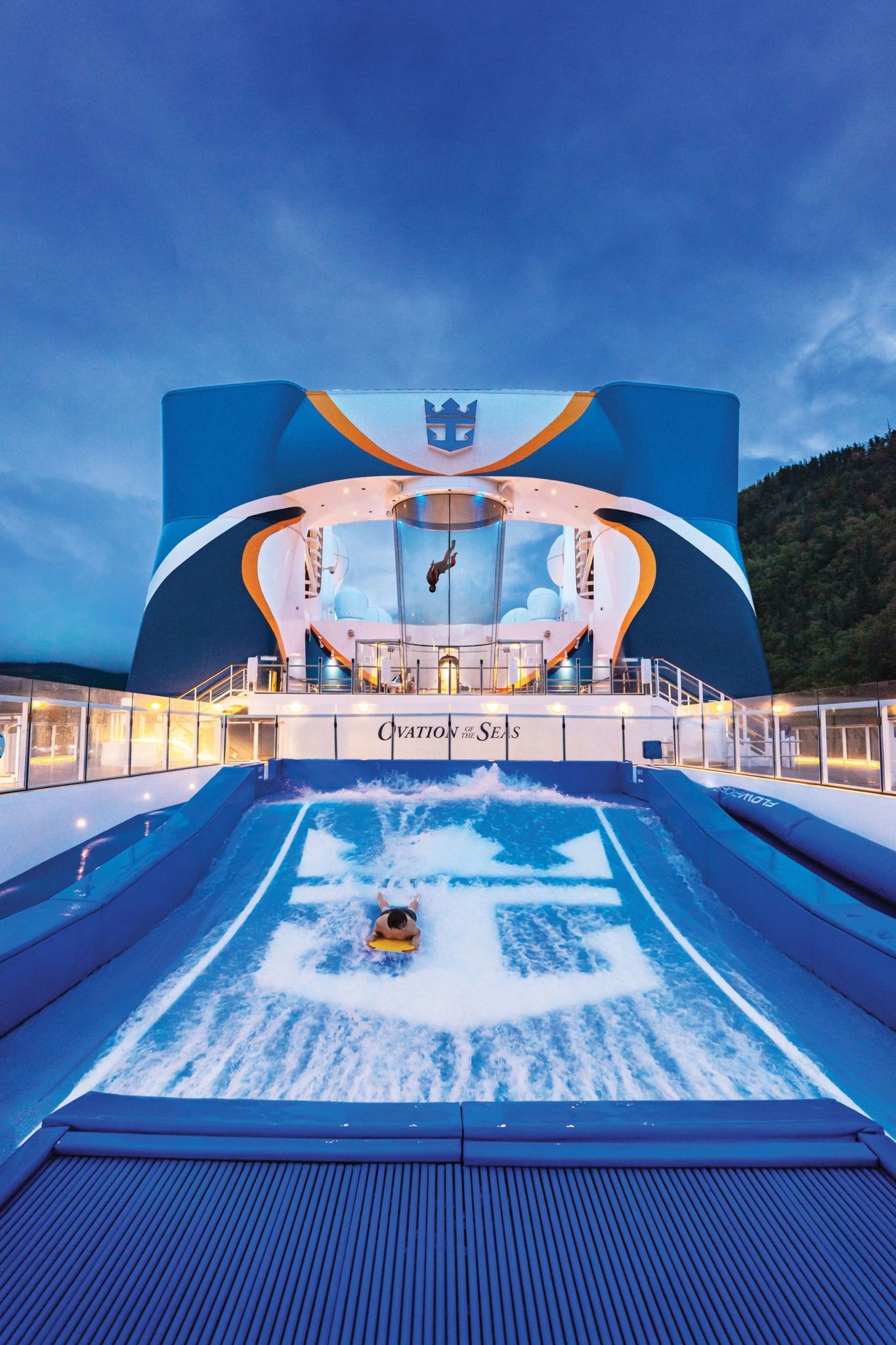 A person surfs on a FlowRider water activity aboard a cruise ship, with a blue and white backdrop under a dusk sky.