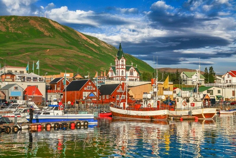 A harbor with colorful buildings and boats, overlooked by a green mountain, under a partly cloudy sky.