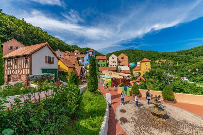 Colorful European-style village nestled in a lush green hillside. People stroll along pathways under a bright blue sky.