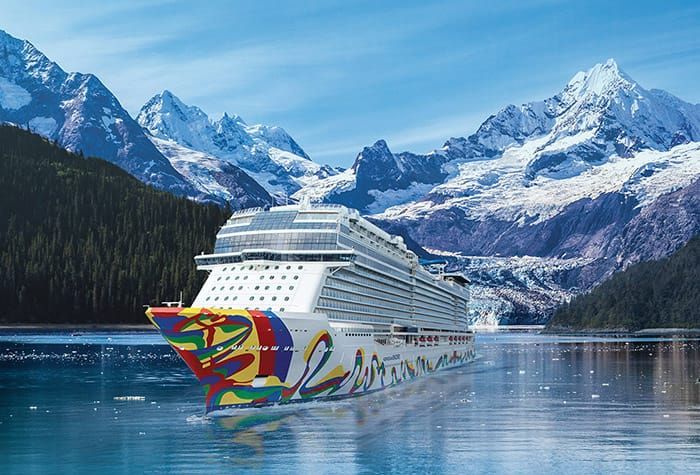 A large cruise ship with colorful art on its bow sails through a glacial fjord, with snowy mountains in the background.