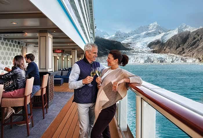 Couple toasting with champagne on a cruise ship deck overlooking a glacier.  Other passengers in the background.