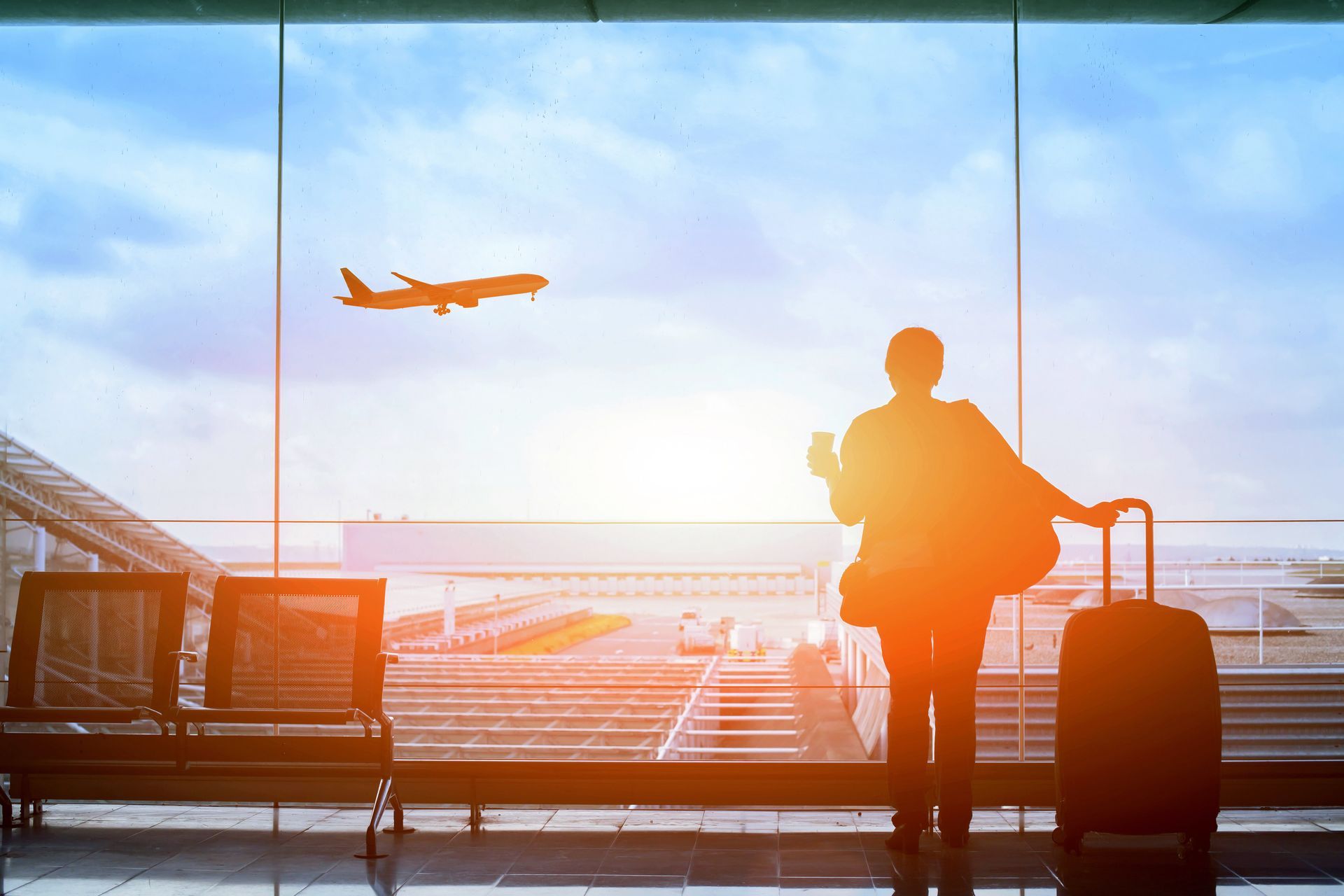 Silhouette of person at airport window, watching plane take off. Luggage beside them, sunlight streaming through glass.