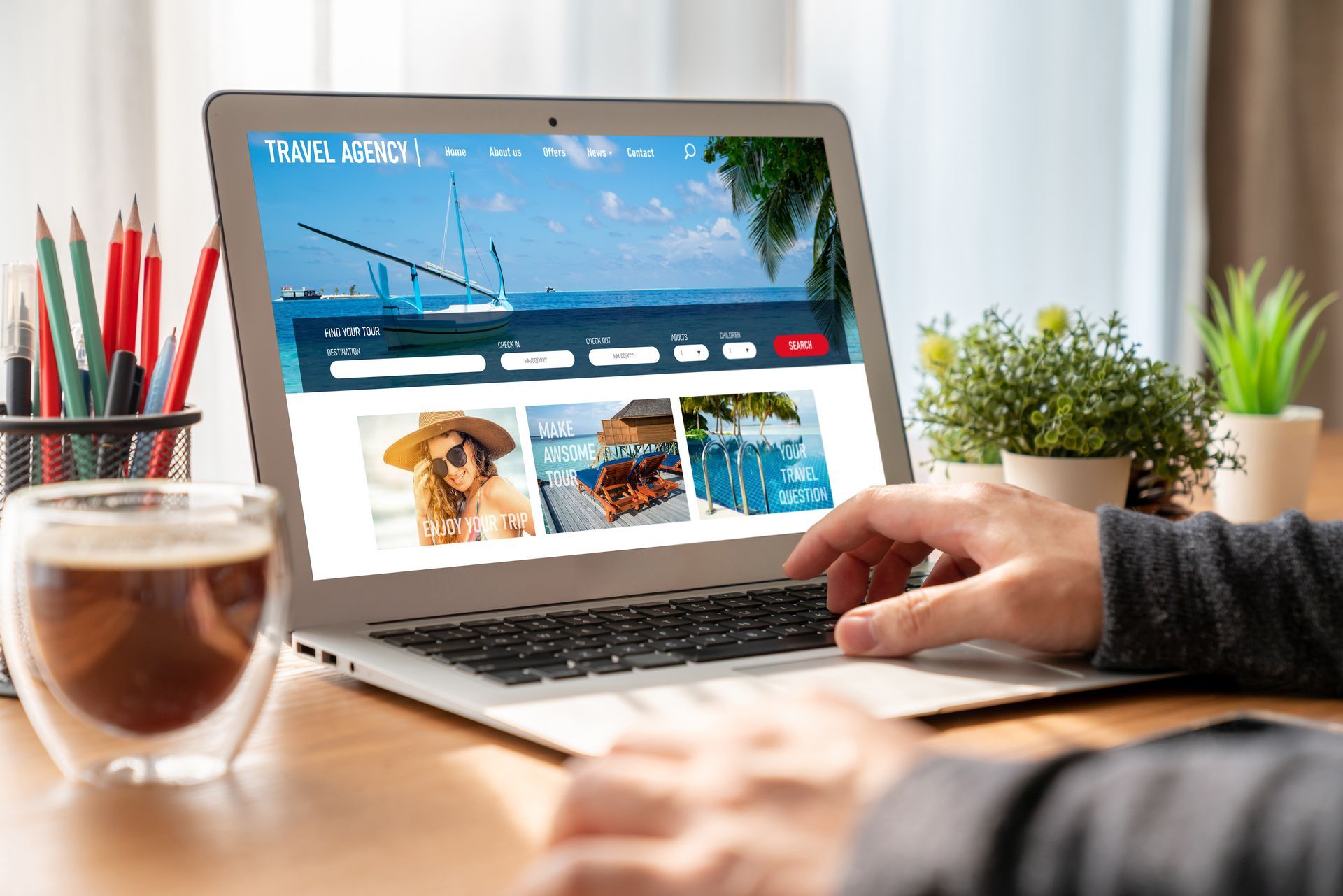 Person using a laptop to browse a travel website, set on a wooden desk with coffee, pens, and plants.