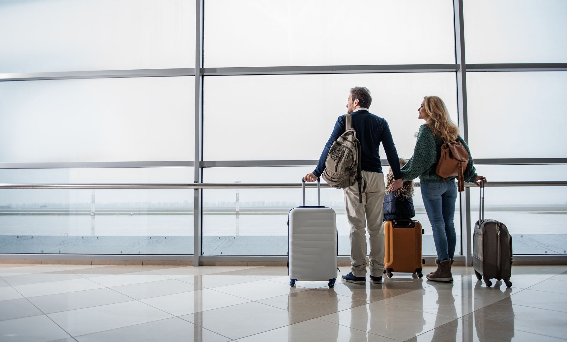 A couple holding hands at an airport, looking out a window. They each have luggage and backpacks, ready for travel.