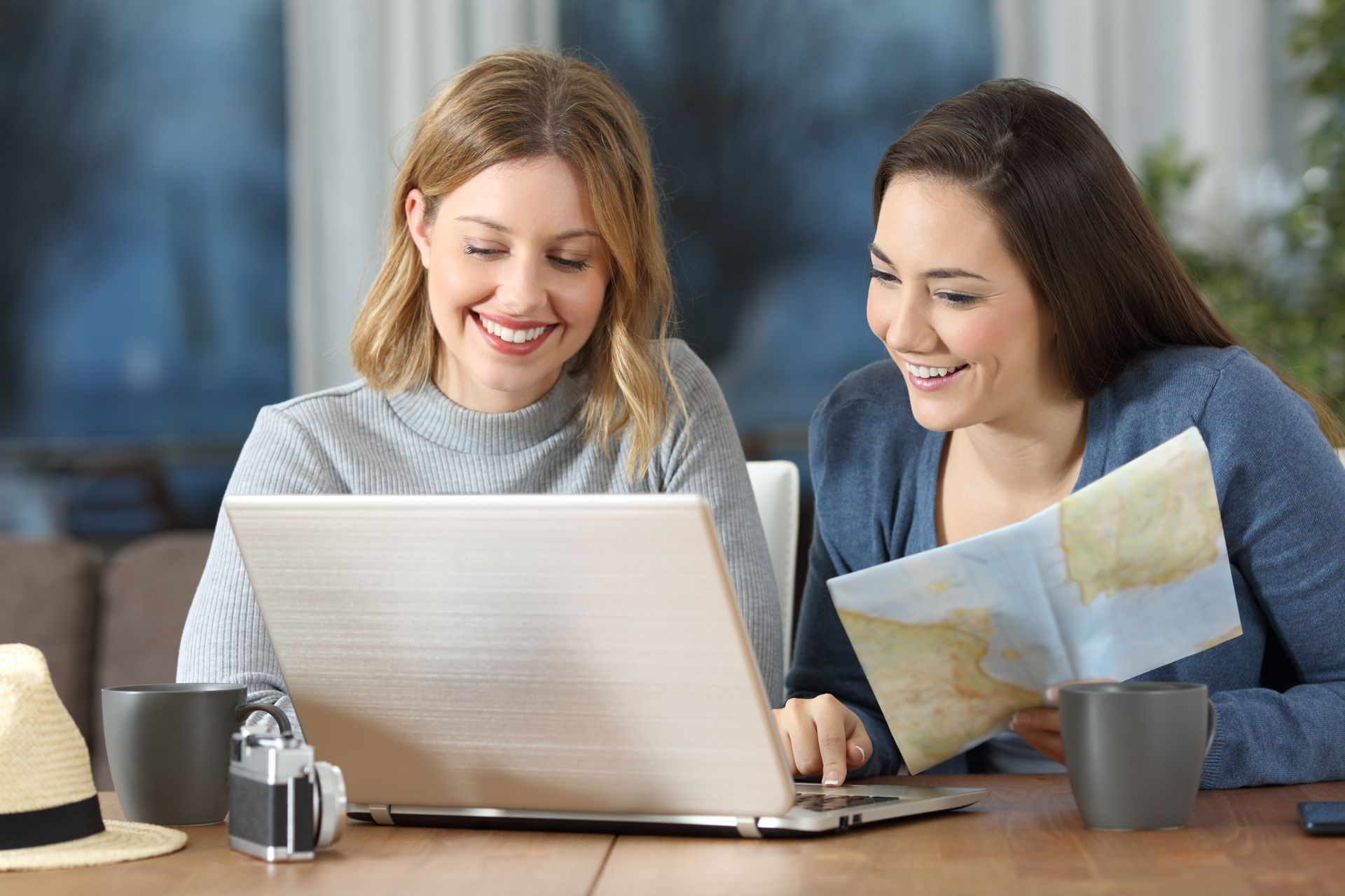 Two women smiling, planning a trip together, one looking at a laptop screen, the other holding a map.