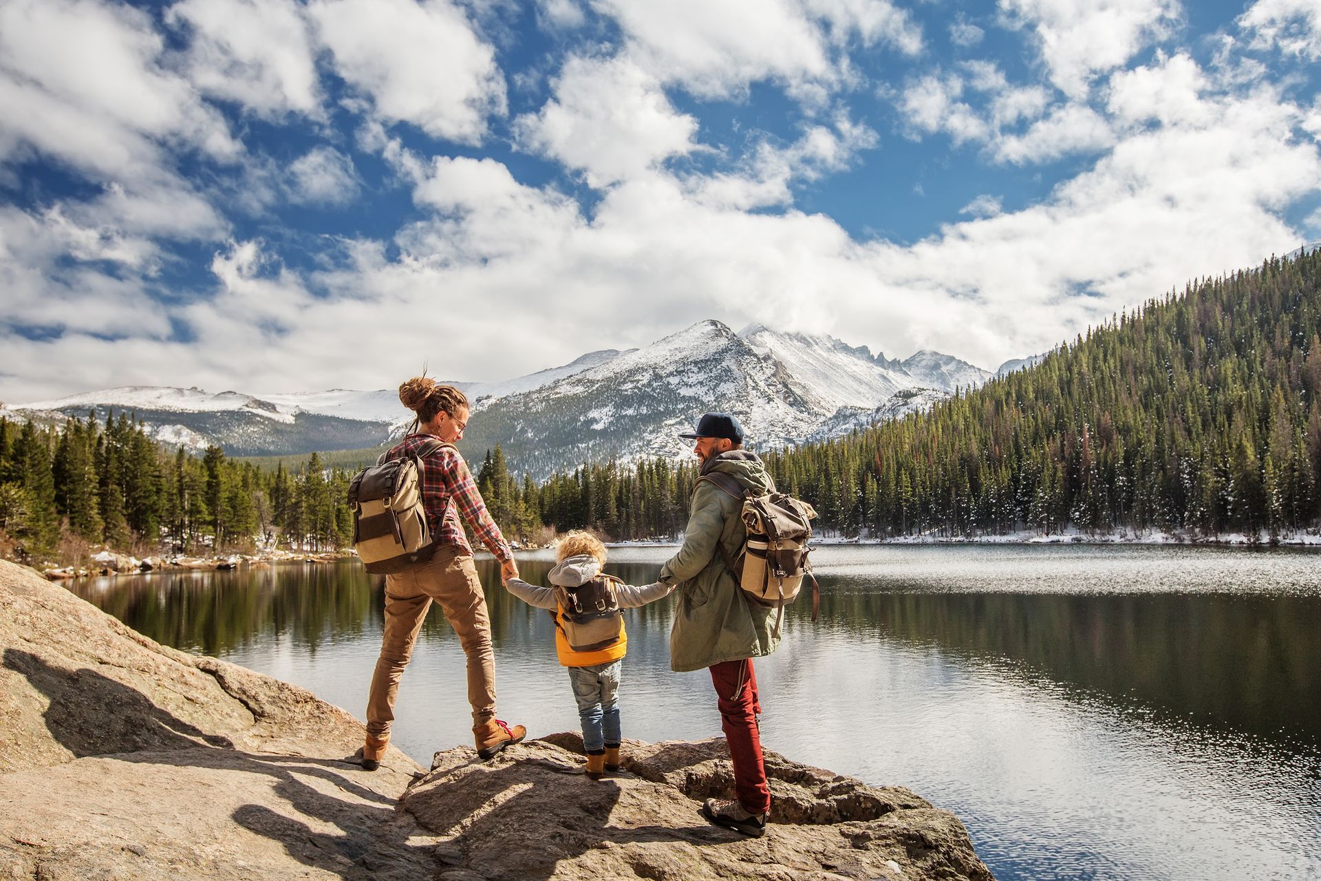 Family of three holding hands, looking out at a lake and snow-capped mountains; sunny day.