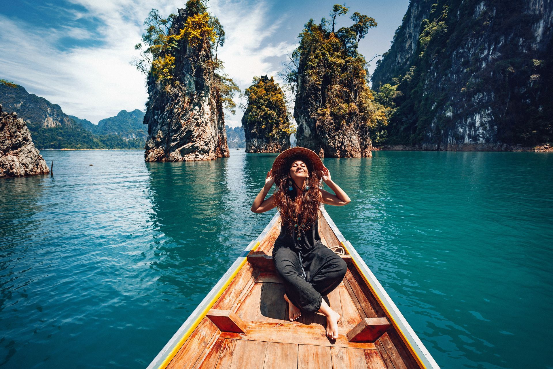Woman in a boat on turquoise water with limestone cliffs in the background, smiling with her arms raised.