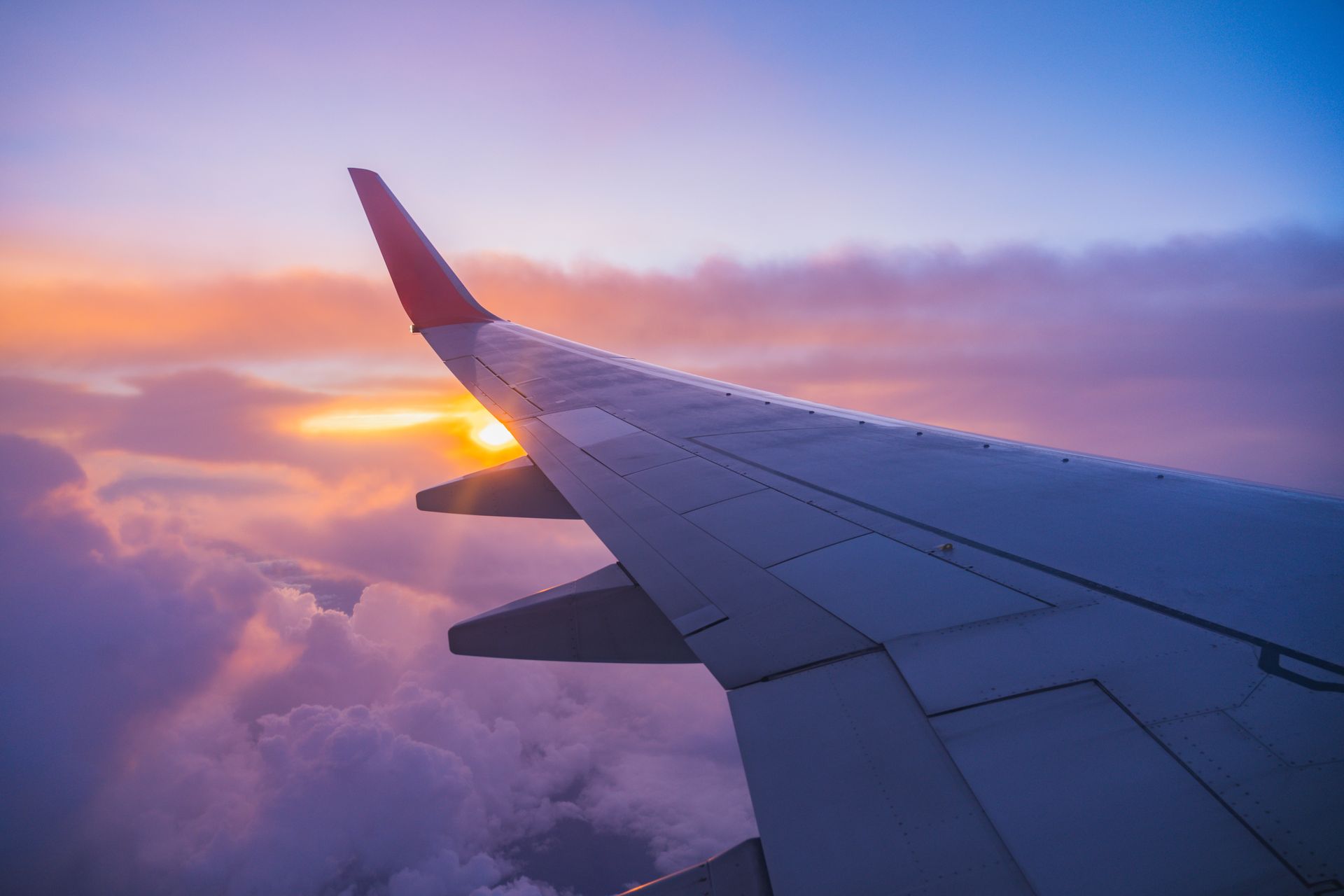 Airplane wing soaring through clouds at sunrise, with orange and purple hues.