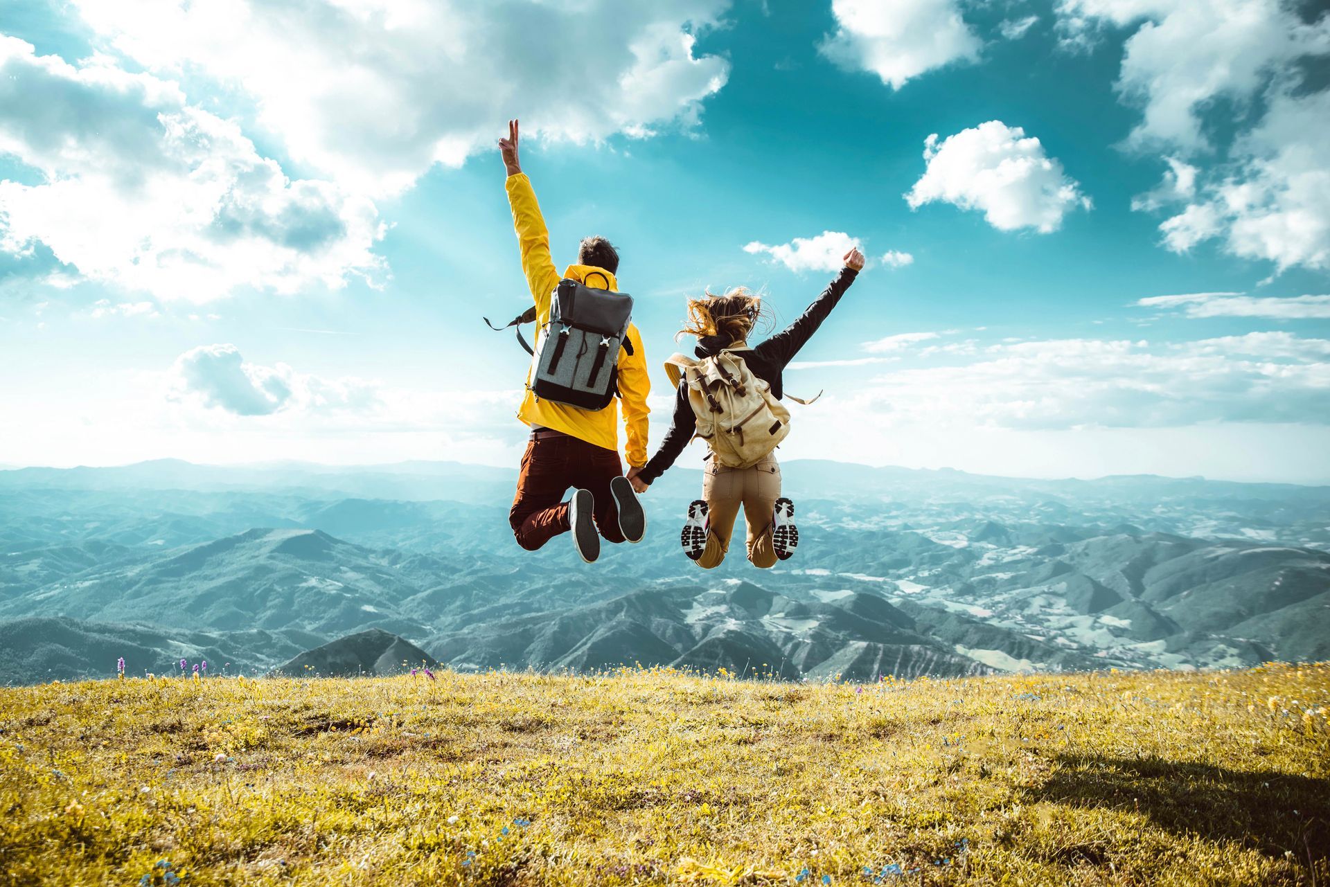 Two hikers jump for joy on a hilltop, arms raised, with a mountainous landscape under a blue sky.