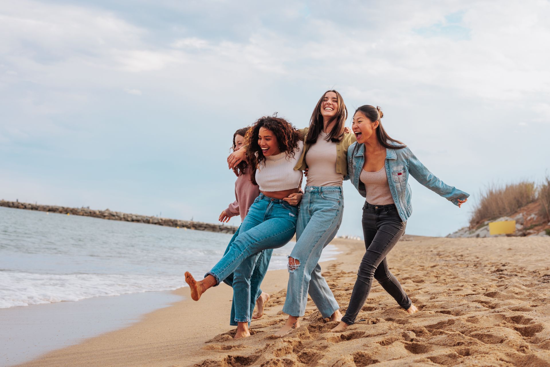 Four young women laughing and walking on a beach, near the water. They are hugging each other and seem joyful on a sunny day.