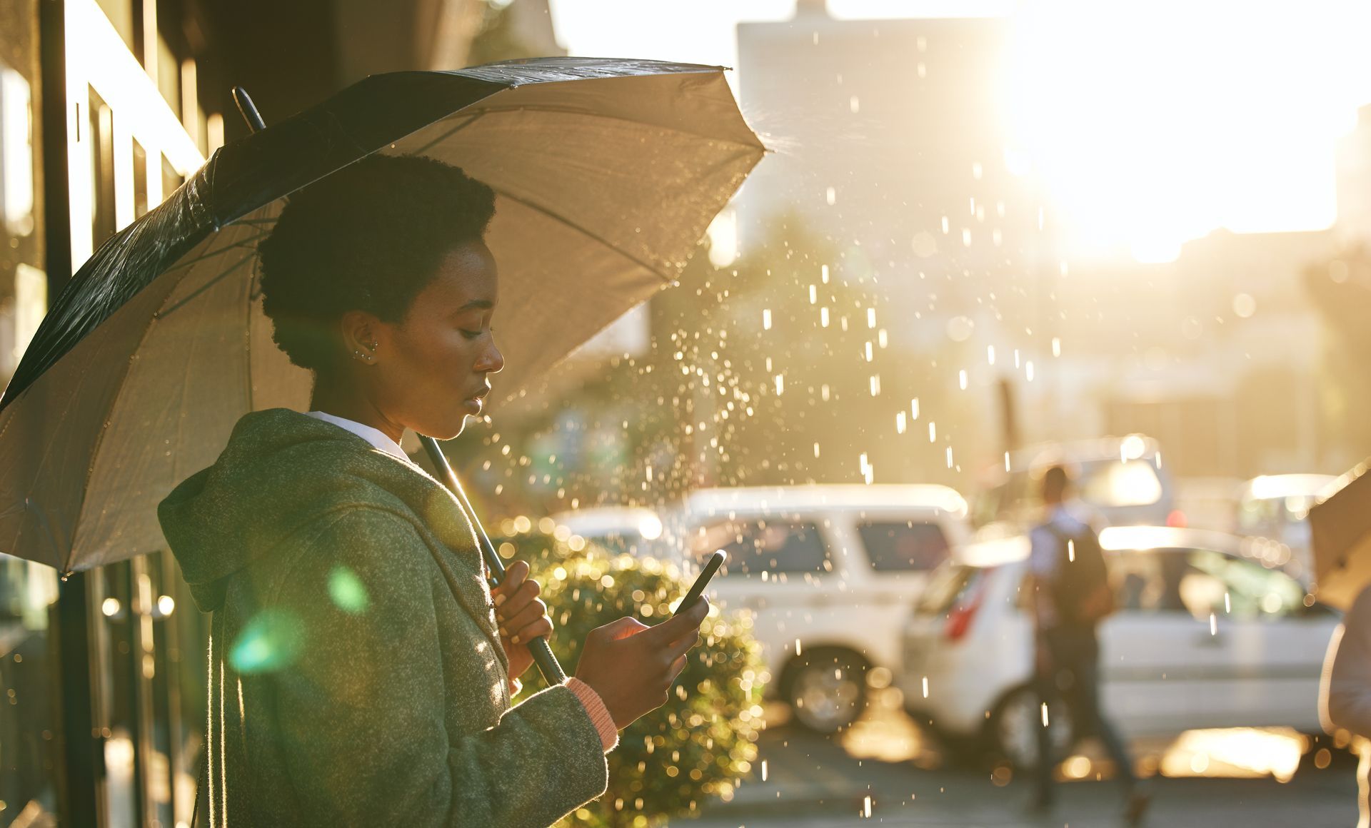 Woman under umbrella in rain, looking at phone. Sunlight shines, illuminating rain and street scene.