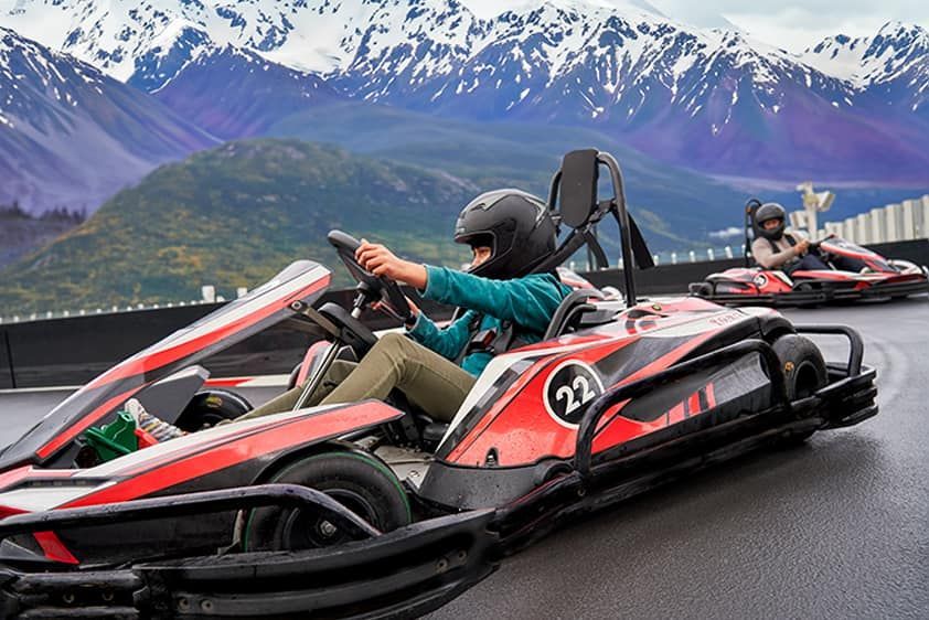 Two people in go-karts race on a track with a mountain backdrop. One driver wears a black helmet and turquoise jacket.